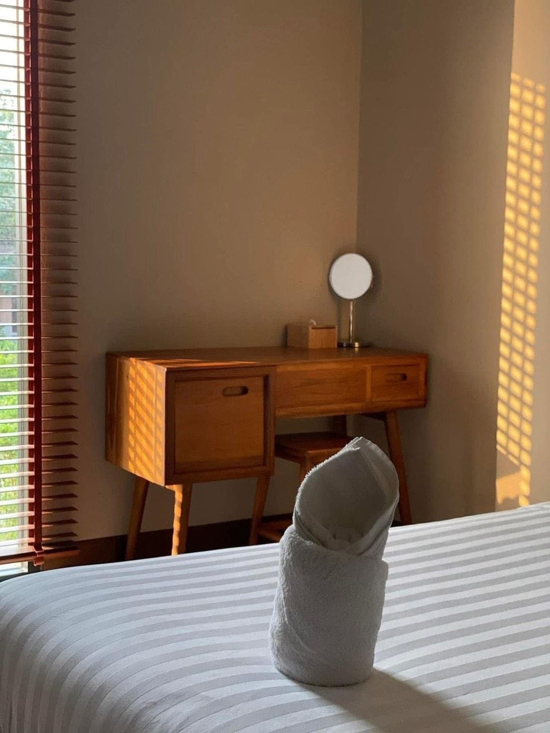 A hotel room with a bed, a wooden desk, a mirror, and a window with blinds.