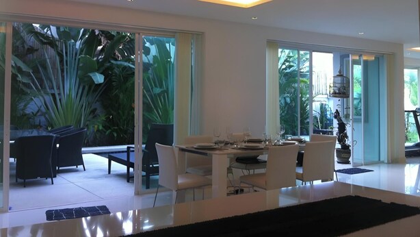 A modern dining area with a glass table, white chairs, and a view of lush greenery outside. - 3-bedroom Pool Villa in lovely Tambon Kamala with cool AC (Tambon Kamala)