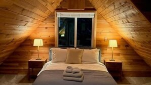 Une chambre confortable située sous les toits, avec un plafond en pente, un grand lit recouvert d’une couvre-lit à rayures, deux lampes de chevet et une fenêtre avec des rideaux.