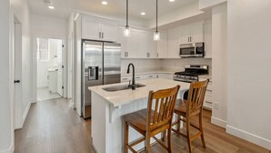 A modern kitchen with white cabinets, stainless steel appliances, and a central island with two wooden chairs. - Foothill Flat | Brand New | Garage | Near Ski Canyons (Sandy)