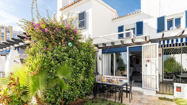 A white building with blue shutters, a patio with a table and chairs, and a lush green garden. - Modern Holiday Home by Beach in Calahonda (Mijas)