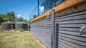 A modern building with a wooden deck, a water feature, and two large tanks in the background. - Piwakawaka Haven - Pohara Beach (Pōhara)