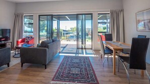 A modern living room with a sofa, a dining table with chairs, a TV, and a large window with curtains. - Piwakawaka Haven - Pohara Beach (Pōhara)