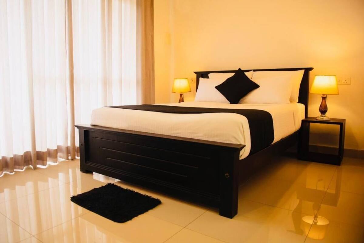 A hotel room with a bed, two bedside lamps, a black rug, and a window with curtains.
