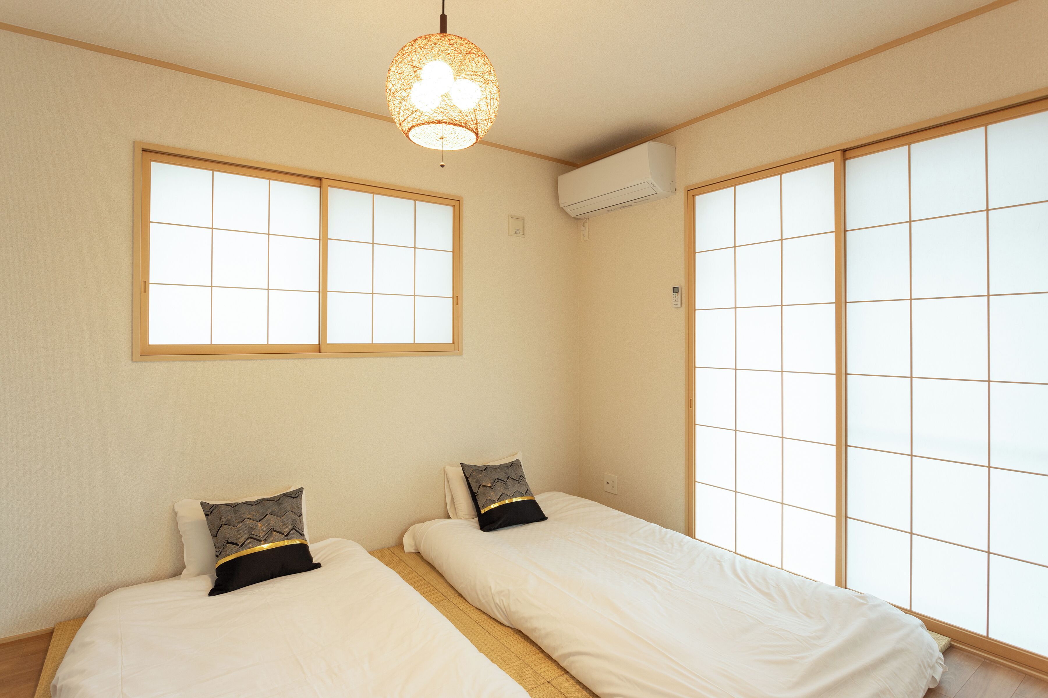 A room with two beds, a large window with grid patterns, an air conditioning unit, and a hanging light fixture.
