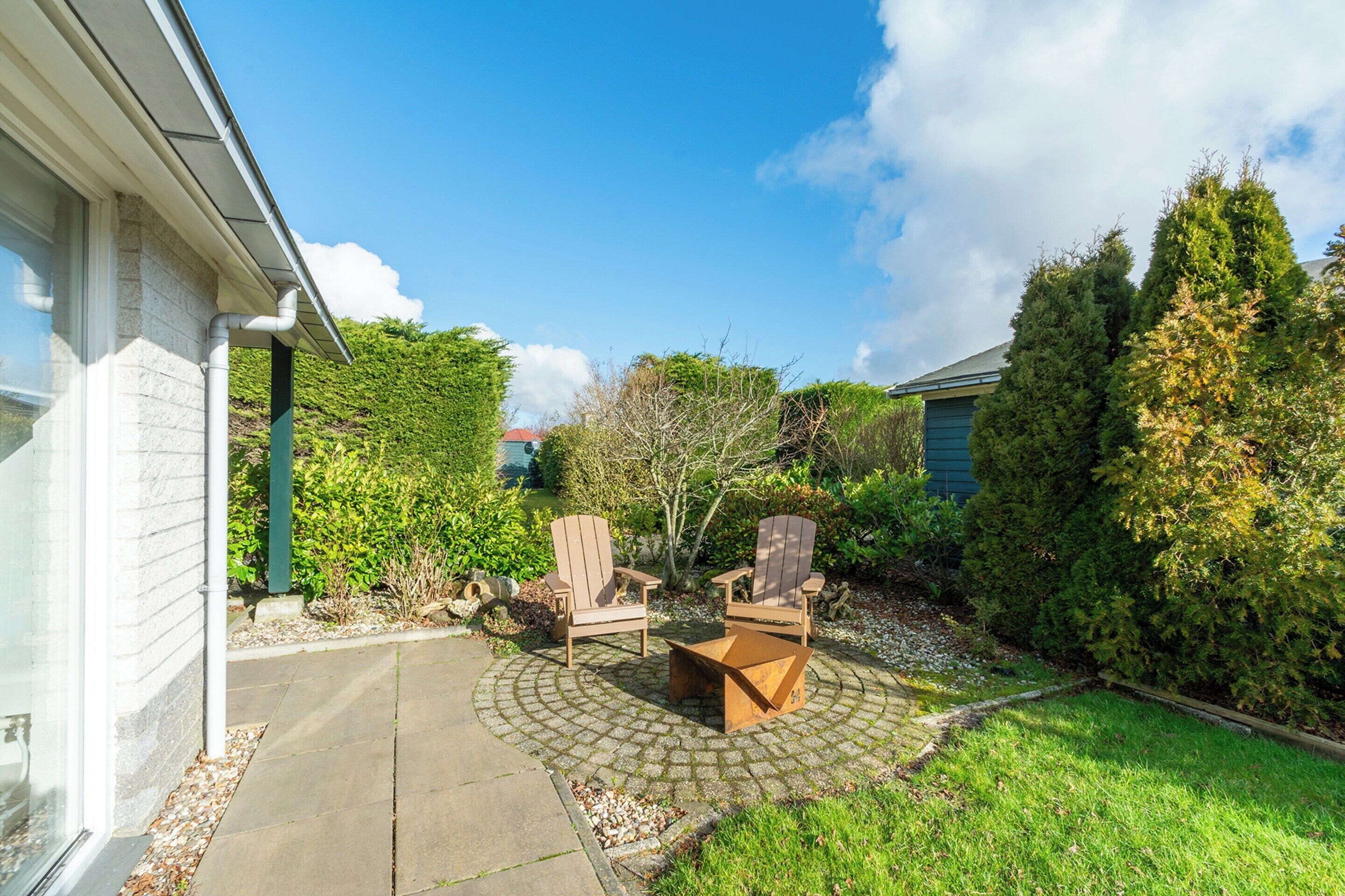 A patio with two Adirondack chairs and a fire pit, surrounded by a hedge and a house.