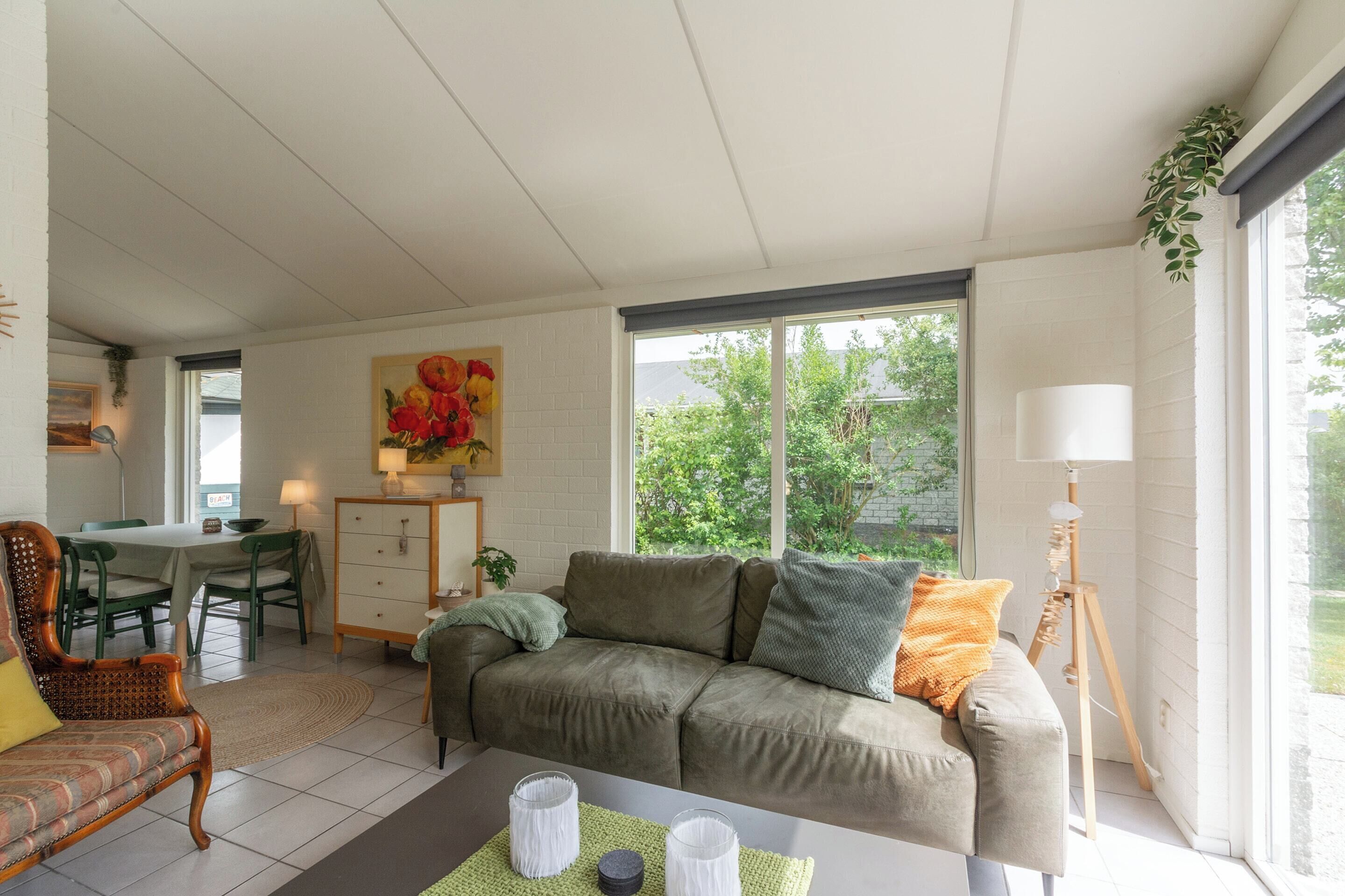 Ein Wohnzimmer mit einer Couch, einem Esstisch und Stühlen. An der Wand hängt ein Gemälde und auf einem Stehlampenfuß steht eine Lampe.