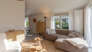 A modern living room with a sofa, a wooden cabinet, and a dining area with a table and chairs. - Holiday Home Near Petten Beach With Pool (Petten)
