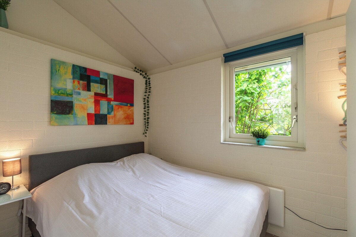A bedroom with a bed, a window with a view of greenery, a wall with a colorful abstract painting, and a bedside table with a lamp.