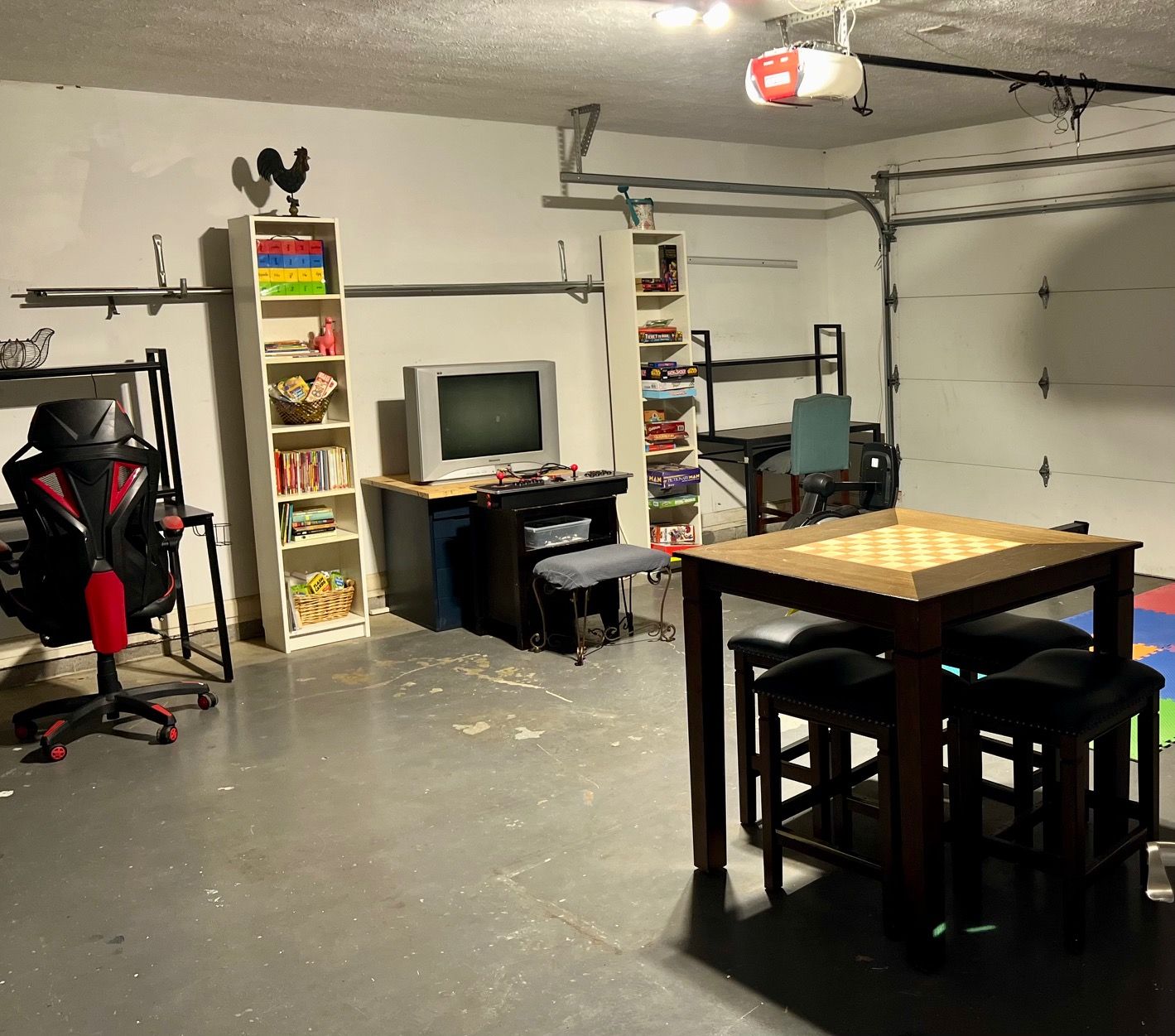 A room with a gaming chair, a desk with a computer, a television, shelves with books and games, and a table with a chessboard.