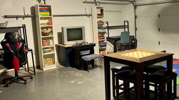 A room with a gaming chair, a desk with a computer, a television, shelves with books and games, and a table with a chessboard.