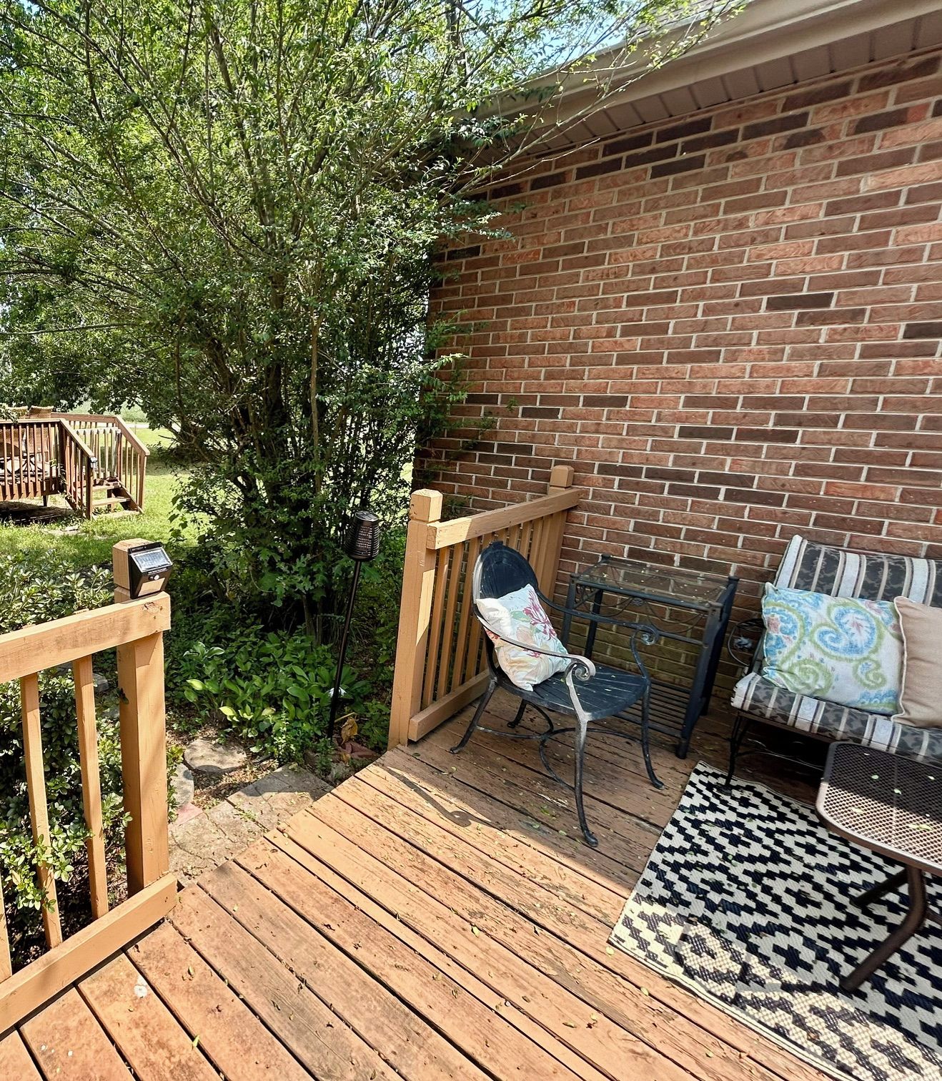 A wooden deck with a metal chair and a cushioned bench, a brick wall, and a small garden area.