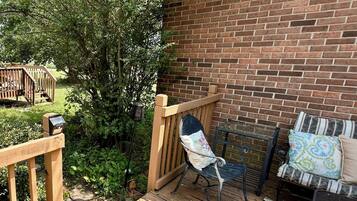 A wooden deck with a metal chair and a cushioned bench, a brick wall, and a small garden area.