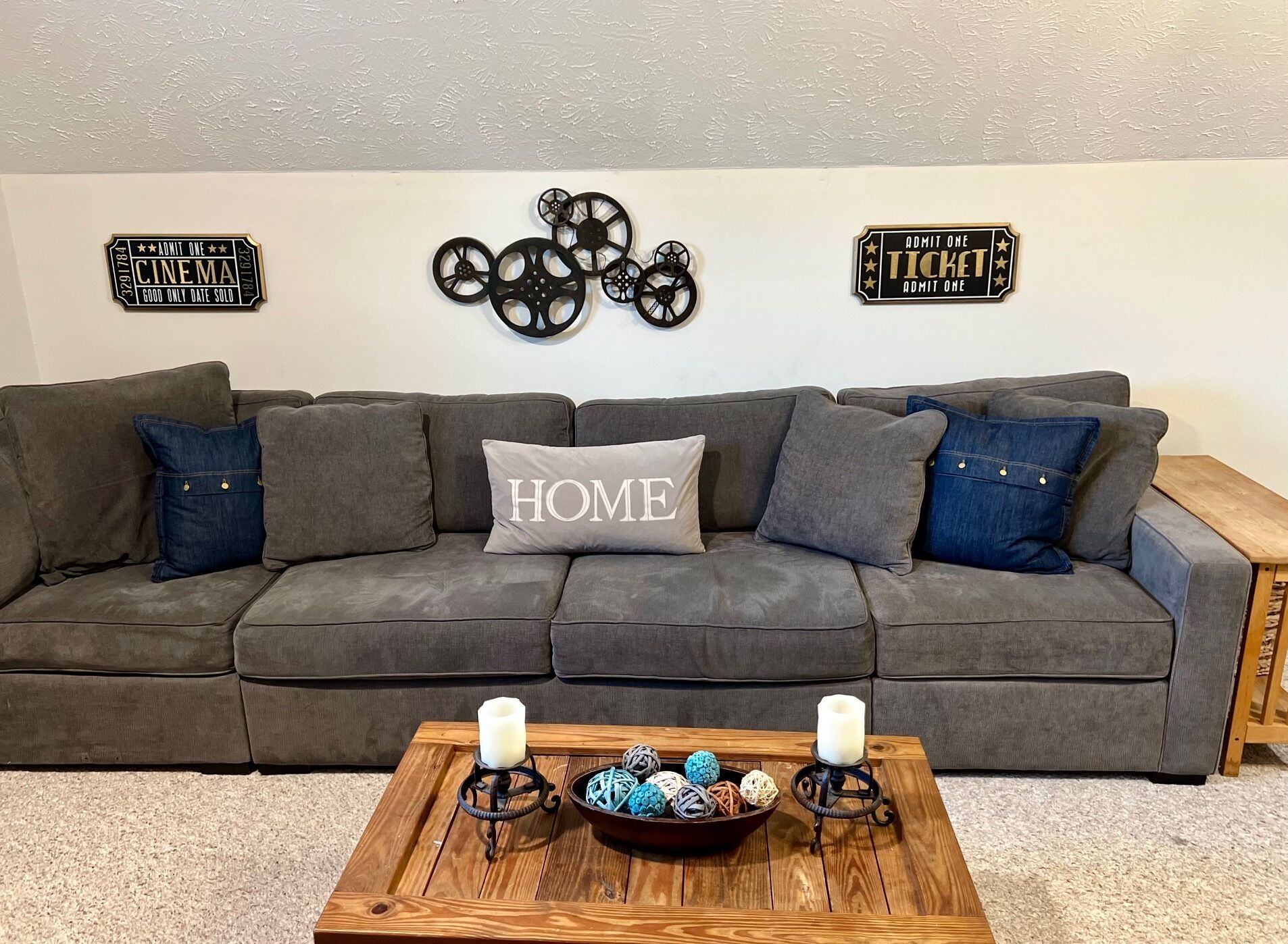 Un canapé d’angle gris avec un coussin orné du mot « HOME », deux panneaux indiquant « UNE PERSONNE », et une table basse en bois sur laquelle sont posées des bougies et des boules décoratives.