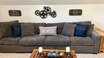 Un canapé d’angle gris avec un coussin orné du mot « HOME », deux panneaux indiquant « UNE PERSONNE », et une table basse en bois sur laquelle sont posées des bougies et des boules décoratives.
