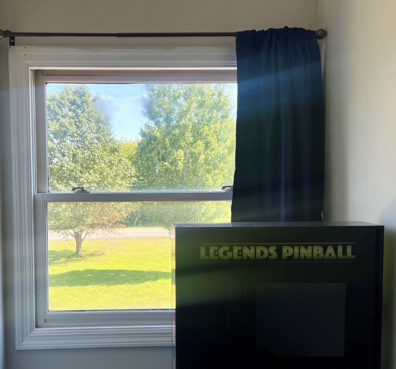 Une fenêtre donnant sur un paysage verdoyant, un rideau et une boîte noire sur laquelle on peut lire le texte « LEGENDS PINBALL ».