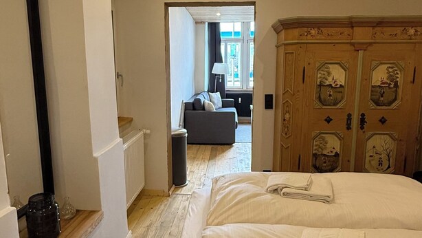 Un dormitorio con piso de madera, una cama con sábanas blancas, un armario de madera con paneles decorativos, y vista a otra habitación con un sofá y una ventana.