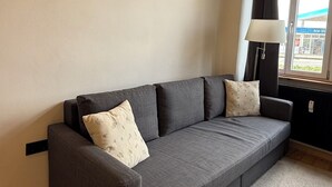 Un sofá gris con cojines estampados, una alfombra blanca, suelo de madera, un techo con vigas de madera y una ventana con vistas a un edificio.