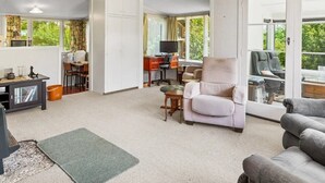 Amplia sala de estar con sofá, sillón y mesa de comedor. Hay un escritorio con computadora y silla. La habitación tiene ventanas grandes y vista a espacios verdes.
