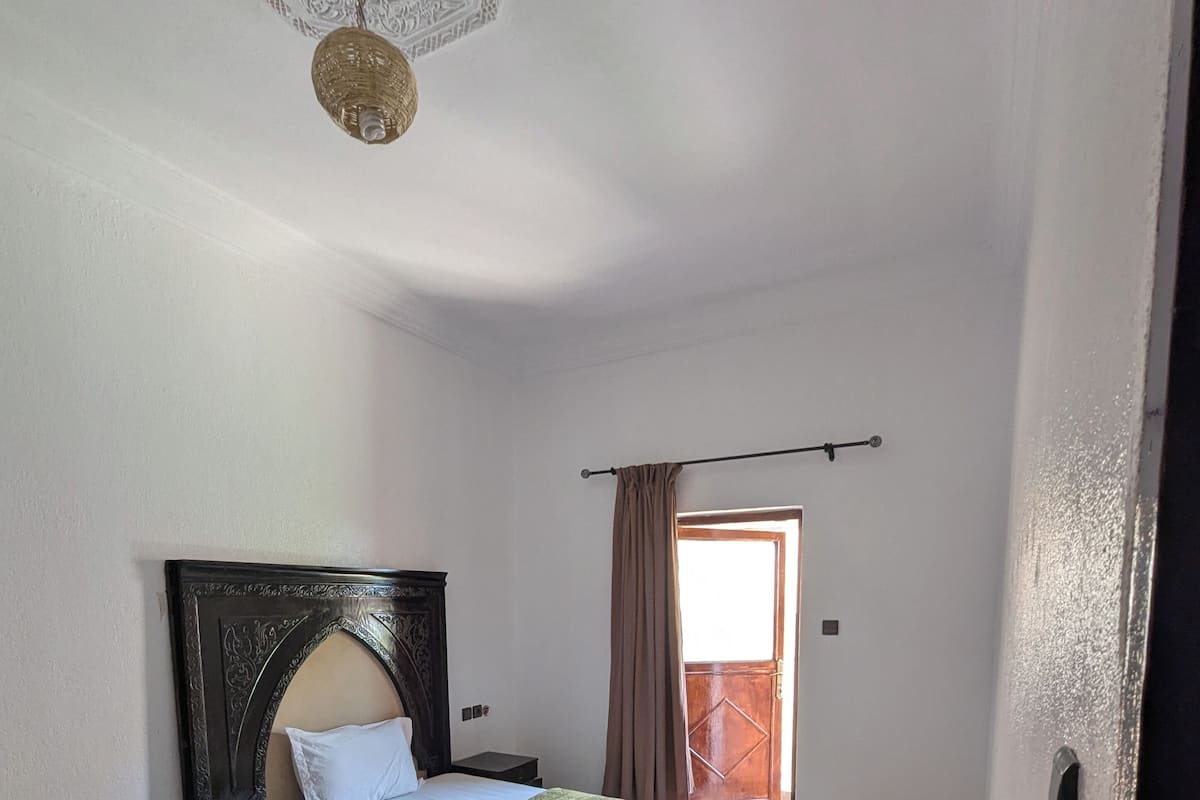 A bedroom with a bed, a nightstand, a red rug, a wooden headboard, and a window with curtains.