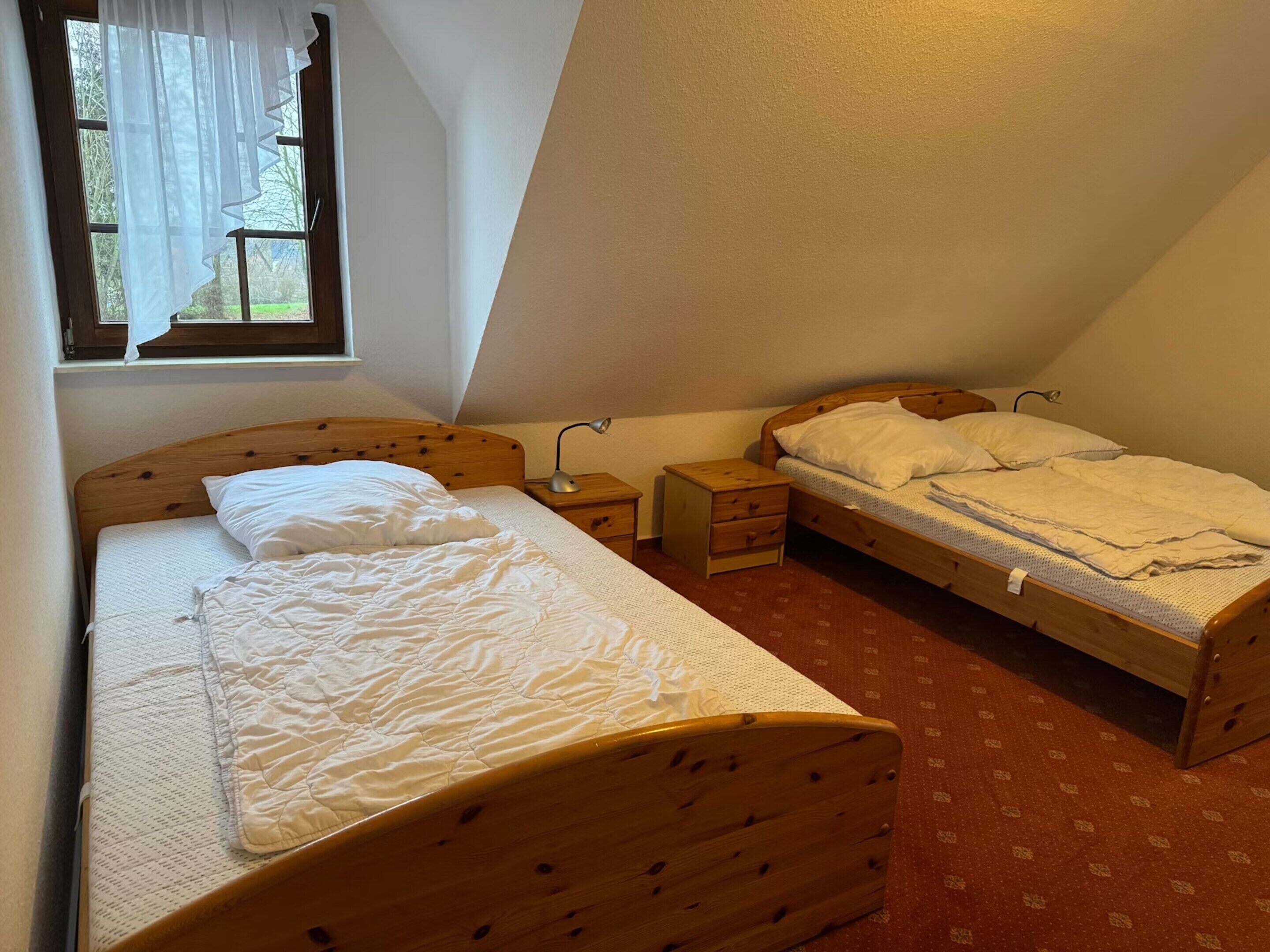 Een slaapkamer met twee houten bedden, een raam met gordijnen, een nachtkastje met een lamp en een vloerbedekking.