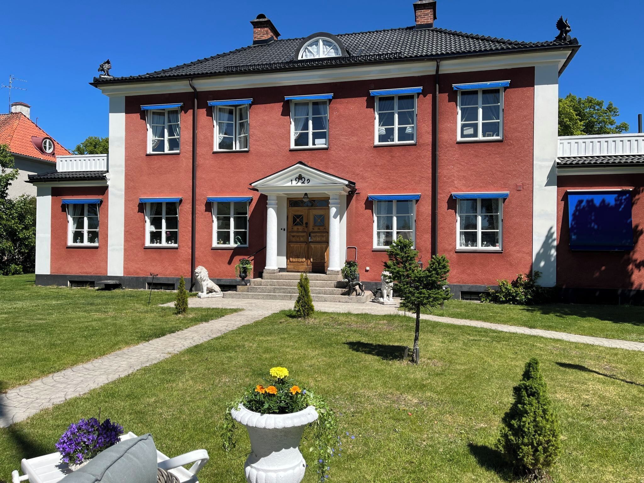 A red brick building with a symmetrical facade, a central entrance with a door, and a small garden with a white bench and flower pots.