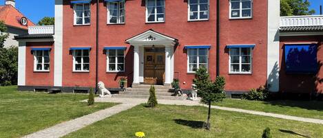 A red brick building with a symmetrical facade, a central entrance with a door, and a small garden with a white bench and flower pots.