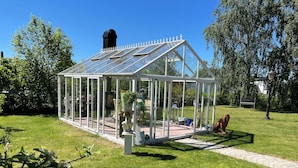 Ein gläsernes Gewächshaus mit Metallverkleidung, umgeben von Rasenfläche und Bäumen.