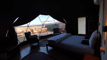 A bedroom with a bed, a desk, and a chair. There is a large window with a view of the outside.