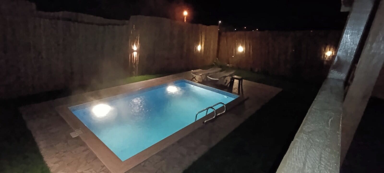A rectangular swimming pool with a ladder, surrounded by a stone wall and a wooden fence.