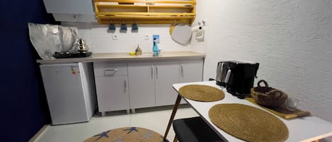 A compact kitchen with a refrigerator, sink, and a coffee maker. There is a dining table with two chairs and a round rug with a star pattern.