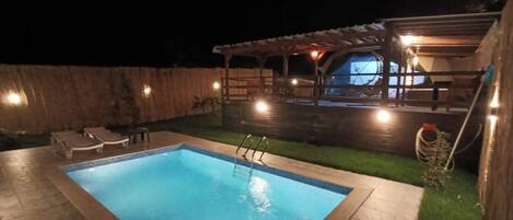 A swimming pool with a wooden deck, lounge chairs, and a covered patio area.