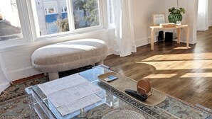 Een kamer met een groot raam, een zachte bank, een glazen tafel met papieren, een afstandsbediening en een klein houten bijzettafeltje met een plant.