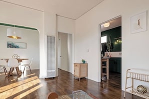 Ein geräumiger Wohnbereich mit Esstisch, Stühlen, einem großen Fenster und einer durchgehenden Tür erkennbaren Küche.