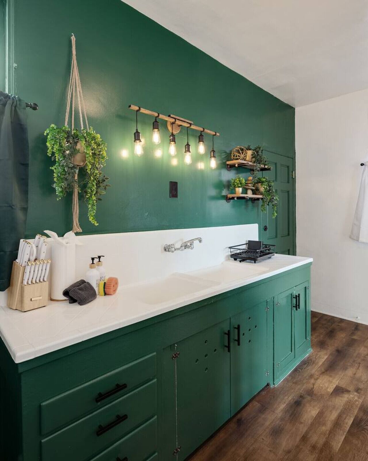Een keuken met een groen kastje, een wit aanrecht, hangende planten en een lamp.
