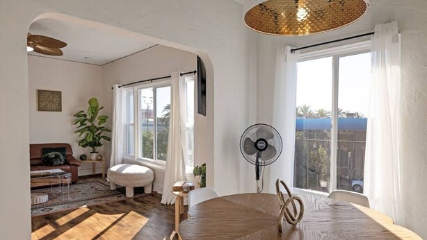 Een ruime woonkamer met een houten eettafel, een staande ventilator en grote ramen die natuurlijk licht binnenlaten.