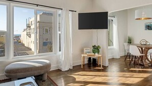 Een moderne woonkamer met een groot raam, een flatscreen-tv, een kleintje tafeltje met een welkomstbord en een eethoek met stoelen.