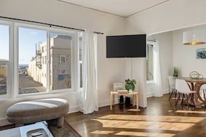 Ein modernes Wohnzimmer mit großem Fenster, einem Flachbildfernseher, einem kleinen Tisch mit einer Willkommensanzeige und einem Essbereich mit Stühlen.