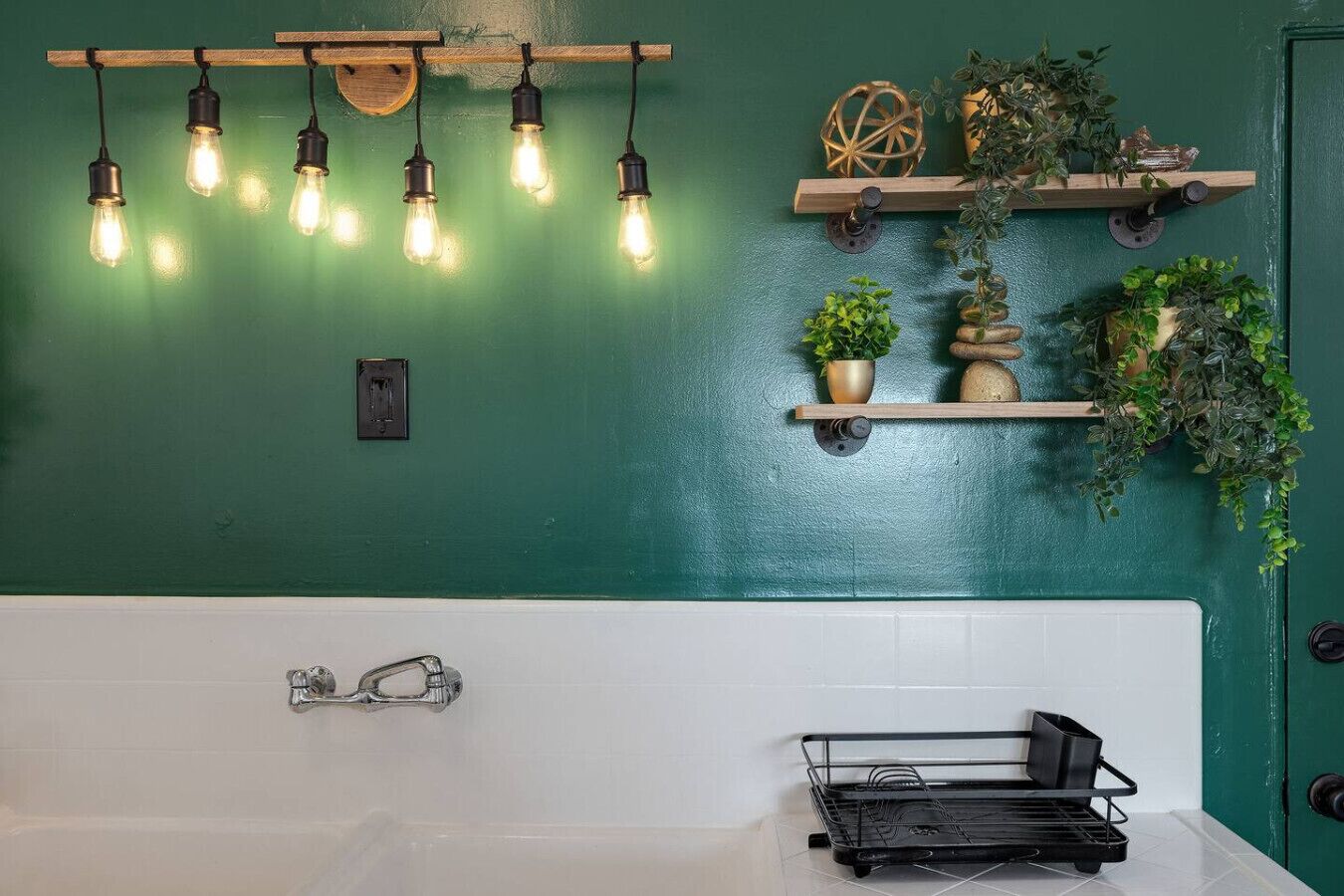 Ein Badezimmer mit einer weißen Badewanne, einem schwarzen Metallregal und einer grünen Wand mit Leuchten und Pflanzenregalen.