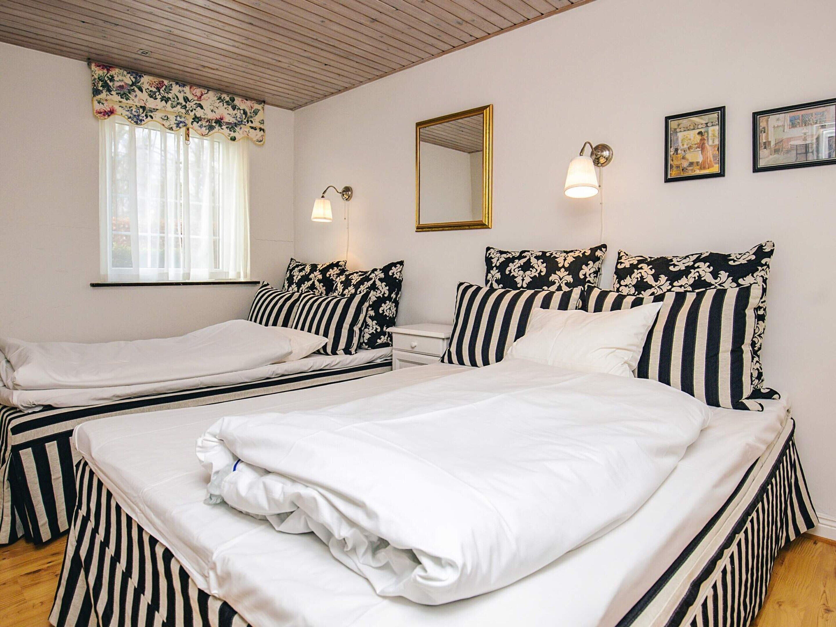 A room with two beds, a window with curtains, a mirror, wall-mounted lights, and framed pictures.