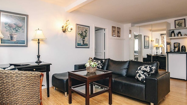 Ein Wohnzimmer mit einer Couch, einem Couchtisch, einem Korbstuhl und einem Schreibtisch mit Lampe. An den Wänden hängen Bilderrahmen, und im Hintergrund ist eine Küchenzeile zu sehen.