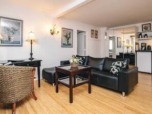 A living room with a sofa, a coffee table, a wicker chair, and a desk with a lamp. There are framed pictures on the walls and a kitchen area in the background. - 4 Person Holiday Home in Brovst (Brovst)