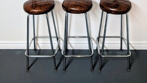 Trois tabourets avec des assises rembourrées et des pieds en métal, une étagère blanche avec une boîte de mouchoirs et des bocaux, et un mur uni.