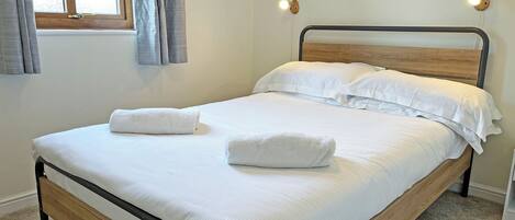 A neatly made bed with white linens and two folded towels, a wooden headboard, a window with curtains, and two wall-mounted lights.