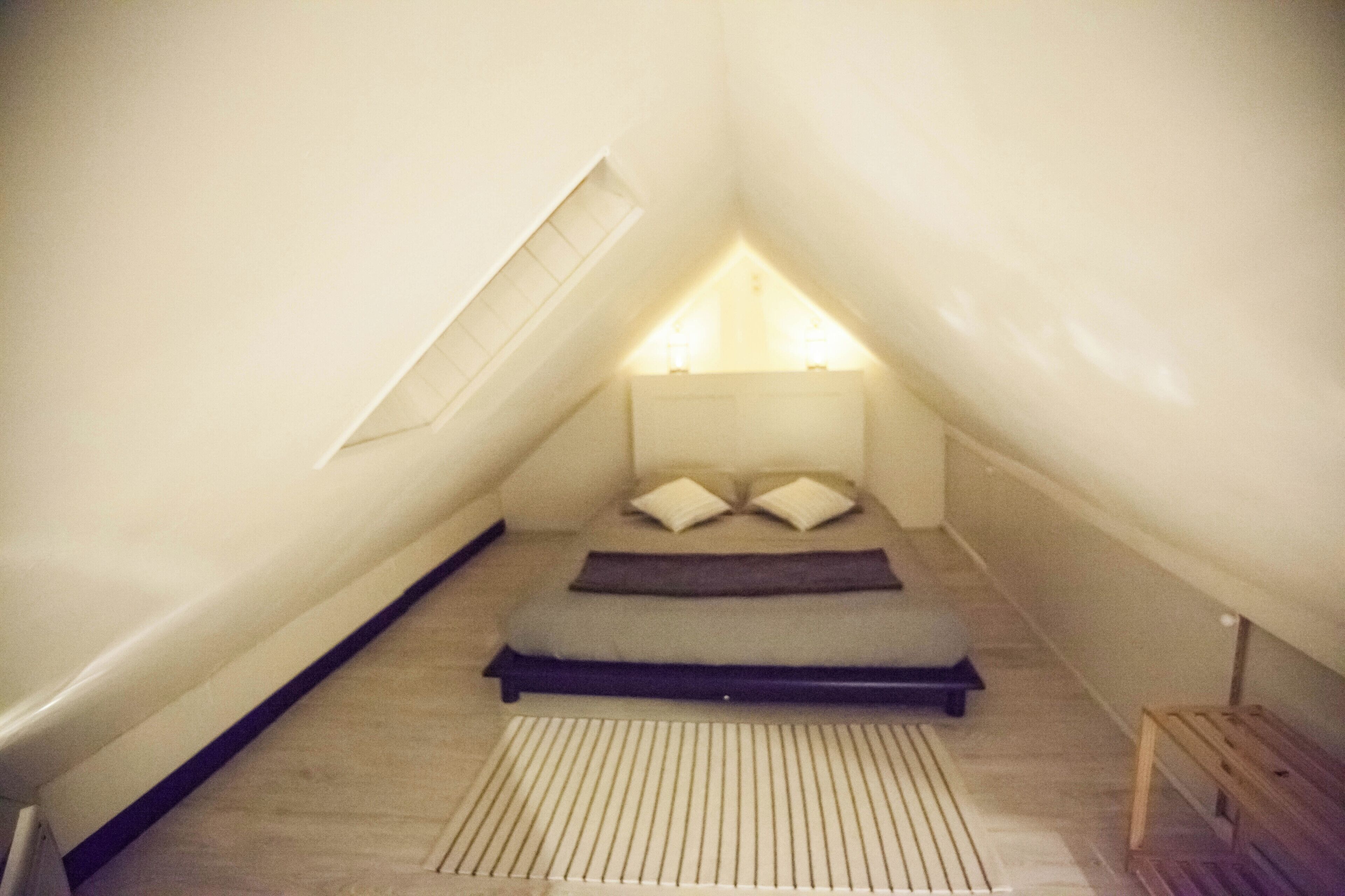 Een kleine, driehoekige slaapkamer onder het dak, met schuine wanden, een bed met kussens en een houten bankje.