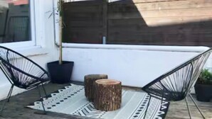 Een houten terras met twee zwarte metalen stoelen, een plant in pot en een klein houten krukje.