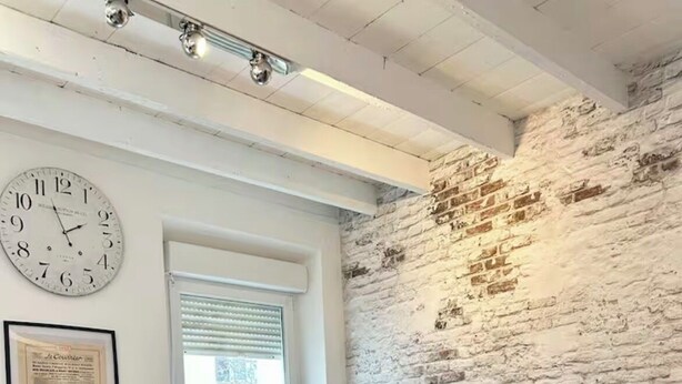 Een woonkamer met een bakstenen muur, een rond zwart poufje, een beige bank met rode kussens, een bijzettafeltje met een lamp en een wandklok.