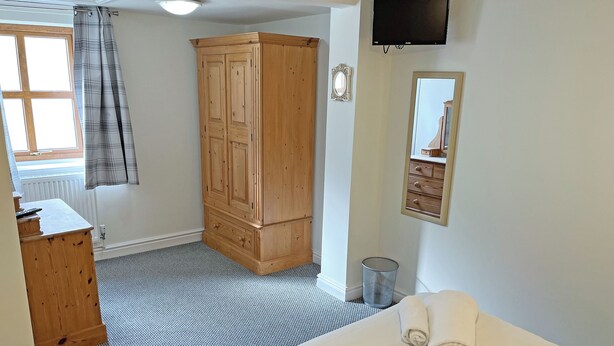 Quarto com cama, guarda-roupa de madeira, televisão, espelho e janela com cortinas.