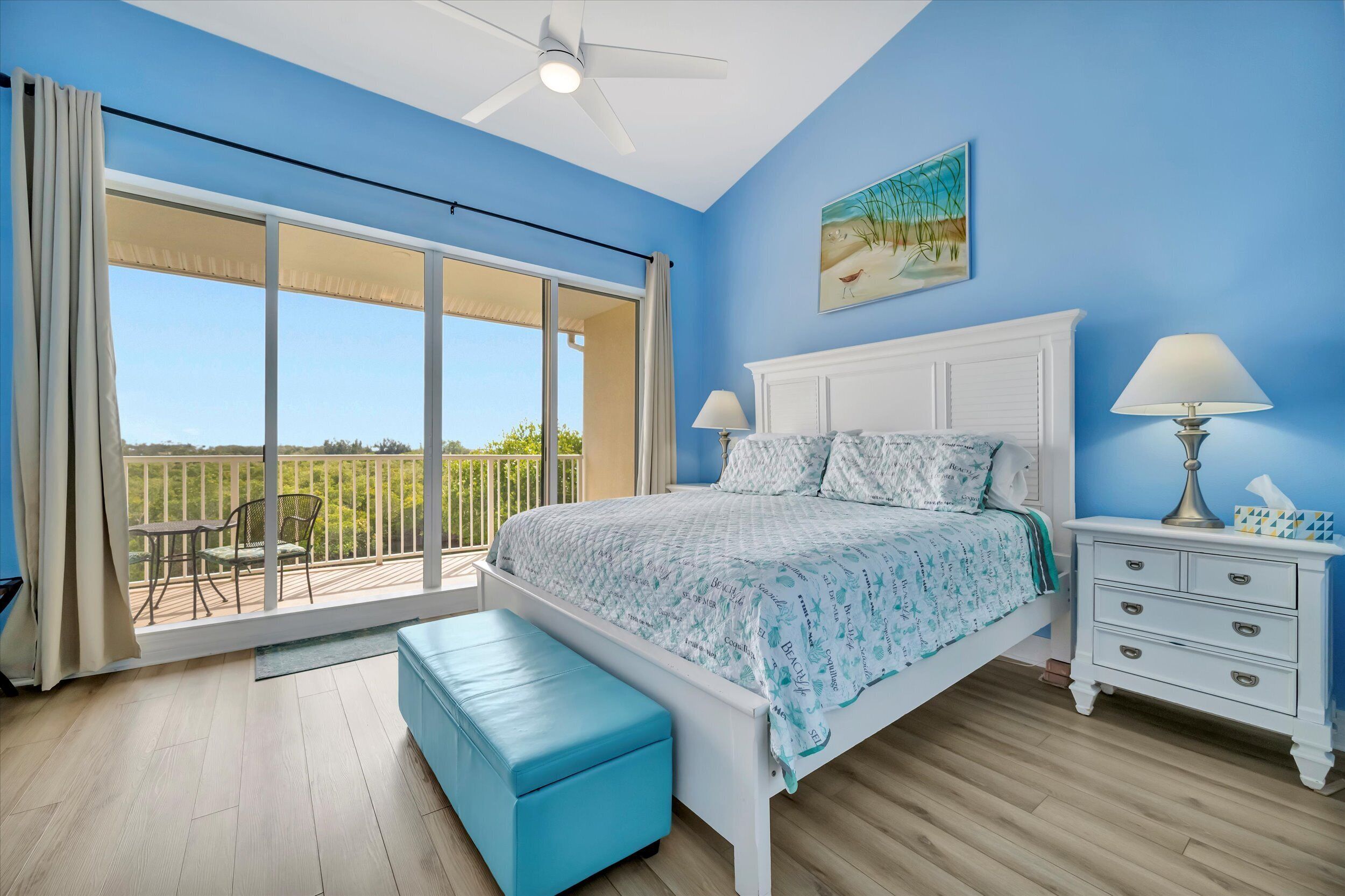 A bedroom with a large sliding glass door to a balcony, a bed with a white headboard, a nightstand with a lamp, and a blue bench.
