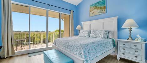 A bedroom with a large sliding glass door to a balcony, a bed with a white headboard, a nightstand with a lamp, and a blue bench.